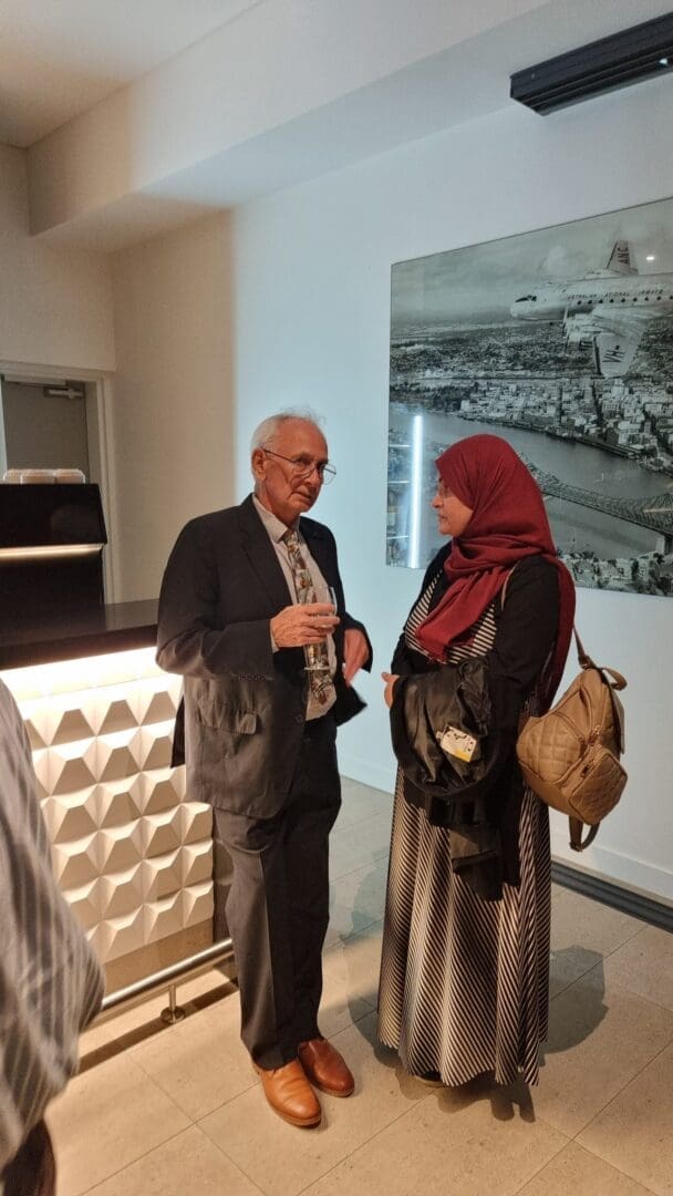 Older white haired man glasses speaking with woman wearing red Hijab large painting behind them OUM