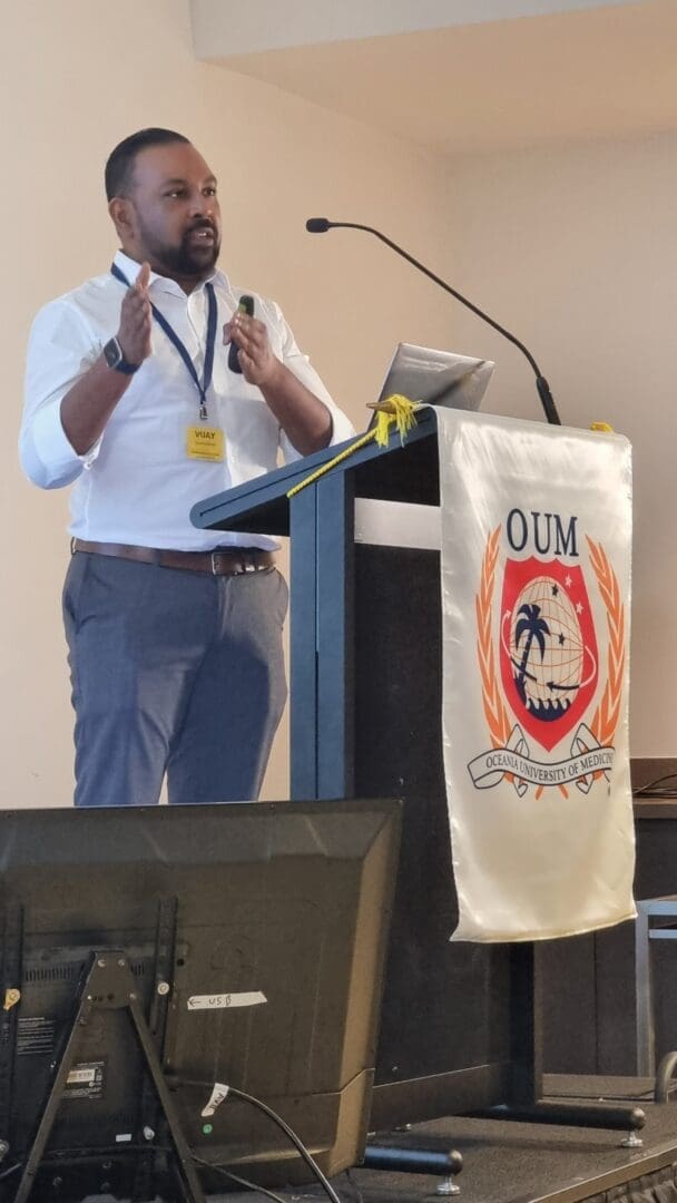 Brown skin man speaking and using his hands by OUM podium banner