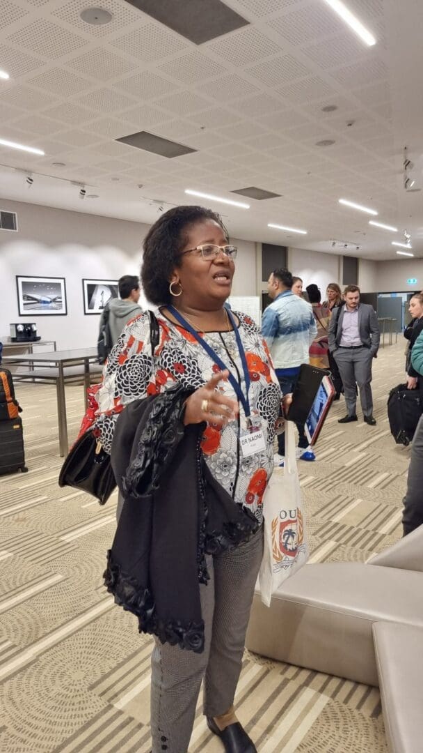 Black woman wire glasses hoop earrings red white shirt black jacket over arm blue lanyard OUM
