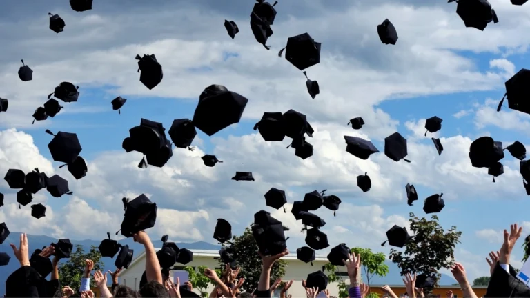 Graduates tossing black caps into the air in celebration against a cloudy blue sky during a graduation ceremony