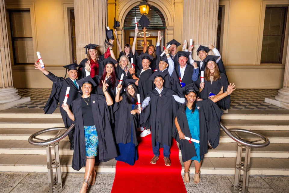 A group of Oceania University of Medicine graduates celebrate together on the steps of a building, wearing black academic gowns and caps, and holding rolled diplomas with red ribbons. They are smiling, raising their hands, and cheering, standing on a red carpet in front of tall columns.
