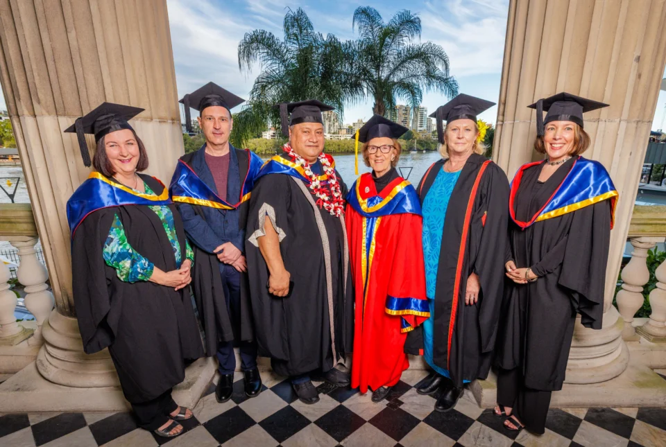 Six staff members in academic regalia pose together between large classical columns. They wear black graduation gowns with colored trim and mortarboards. The person in the center wears a distinctive red and blue ceremonial robe.