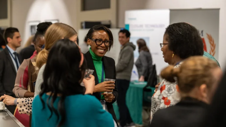 Group of professionals networking and conversing at a conference, with a smiling woman in glasses and a green turtleneck holding a drink at the center
