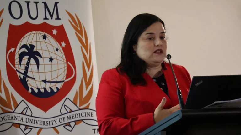 Speaker in a red blazer presenting at a podium with a laptop, standing beside an Oceania University of Medicine banner