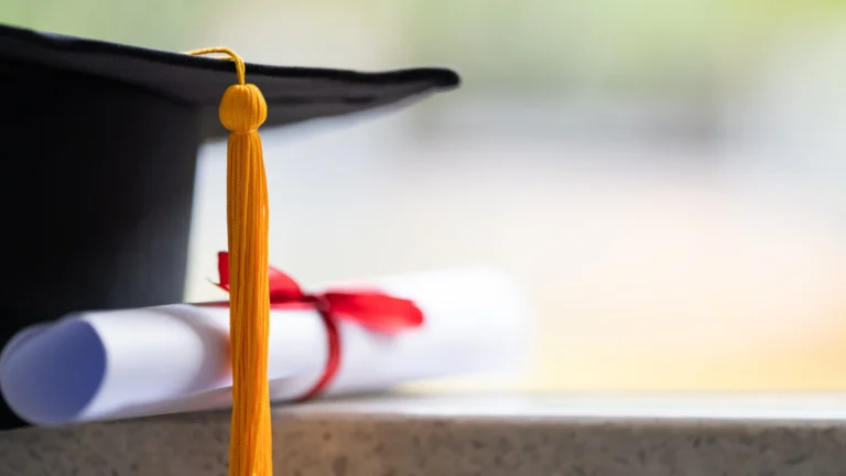 Graduation cap with a yellow tassel resting next to a rolled diploma tied with a red ribbon