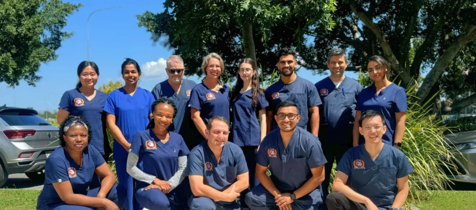 A team healthcare professionals, men and women of diverse backgrounds, wearing matching dark blue scrubs with logos, posed together outdoors under large shade trees.