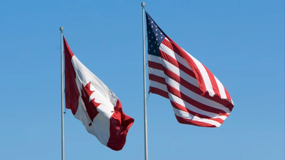 Two flags, the Canadian and the US, waving in the wind against a clear blue sky.