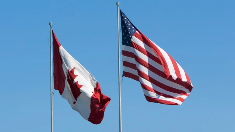 Two flags, the Canadian and the US, waving in the wind against a clear blue sky.