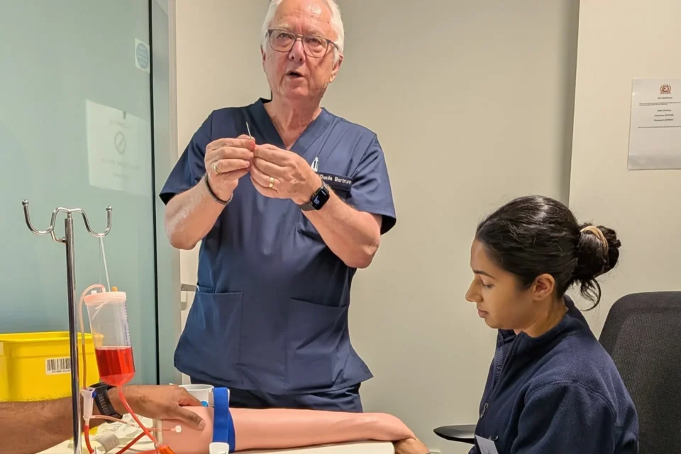 A medical professional in blue scrubs is demonstrating a blood-drawing technique to a younger colleague in a medical setting.