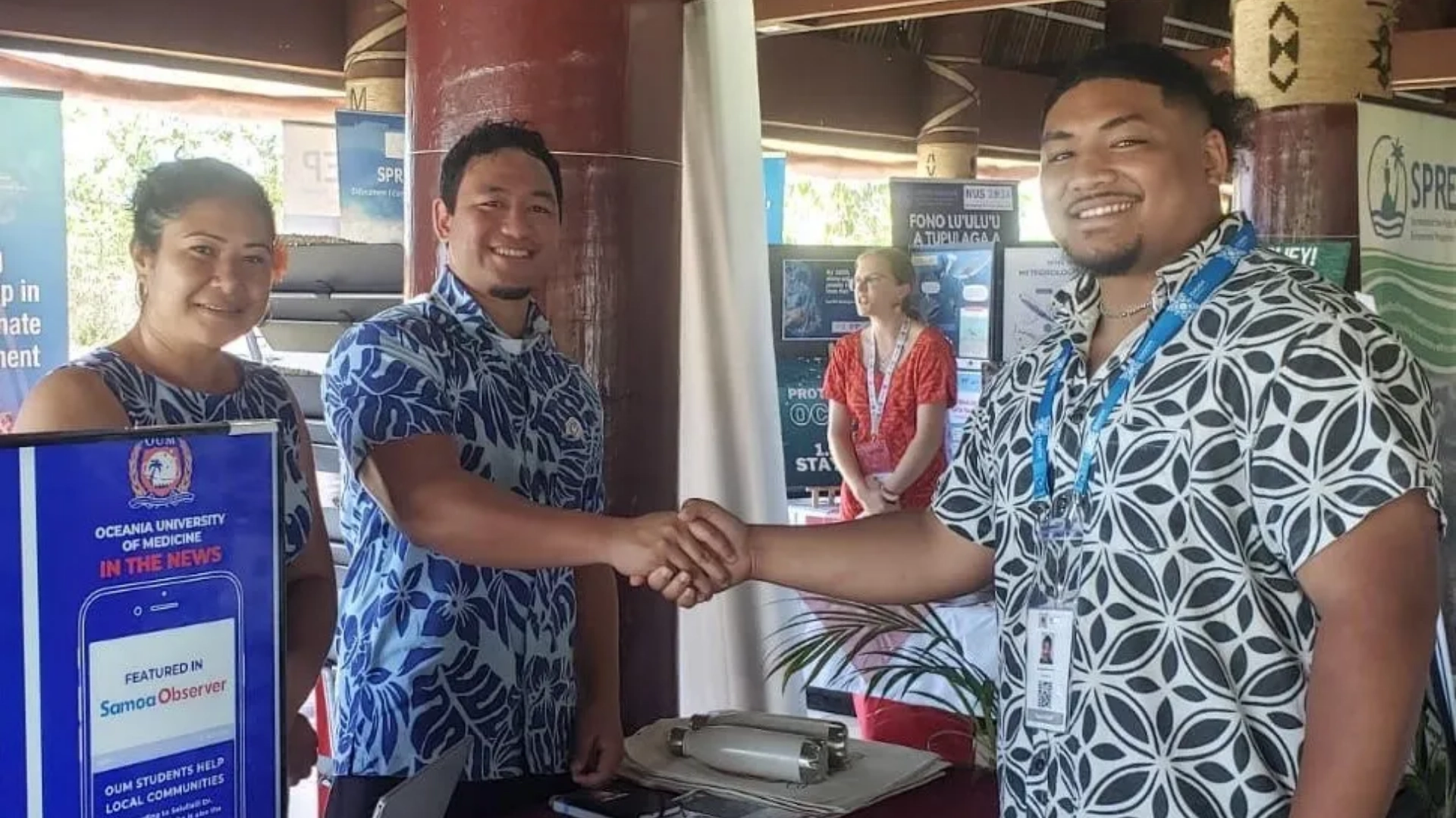 Two men shaking hands and smiling at an Oceania University of Medicine booth during a CHOGM event in Samoa, with a woman and informational displays in the background