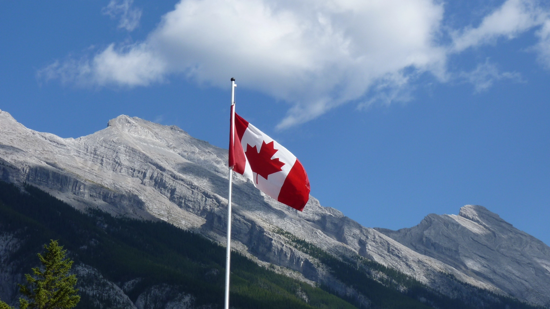 Canadian flag waving on a pole with rocky mountains and a bright blue sky in the background