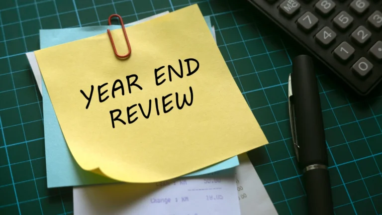 Yellow sticky note with the words “Year End Review” clipped to papers on a desk, with a calculator and pen nearby