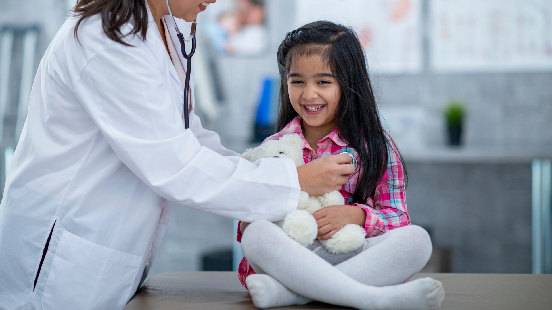 Smiling young girl holding a stuffed animal while a doctor in a white coat uses a stethoscope to listen to her chest during a pediatric checkup