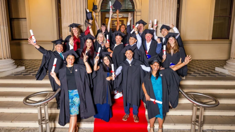 Group of graduates in caps and gowns celebrating on a staircase with diplomas in hand, smiling and cheering after a graduation ceremony