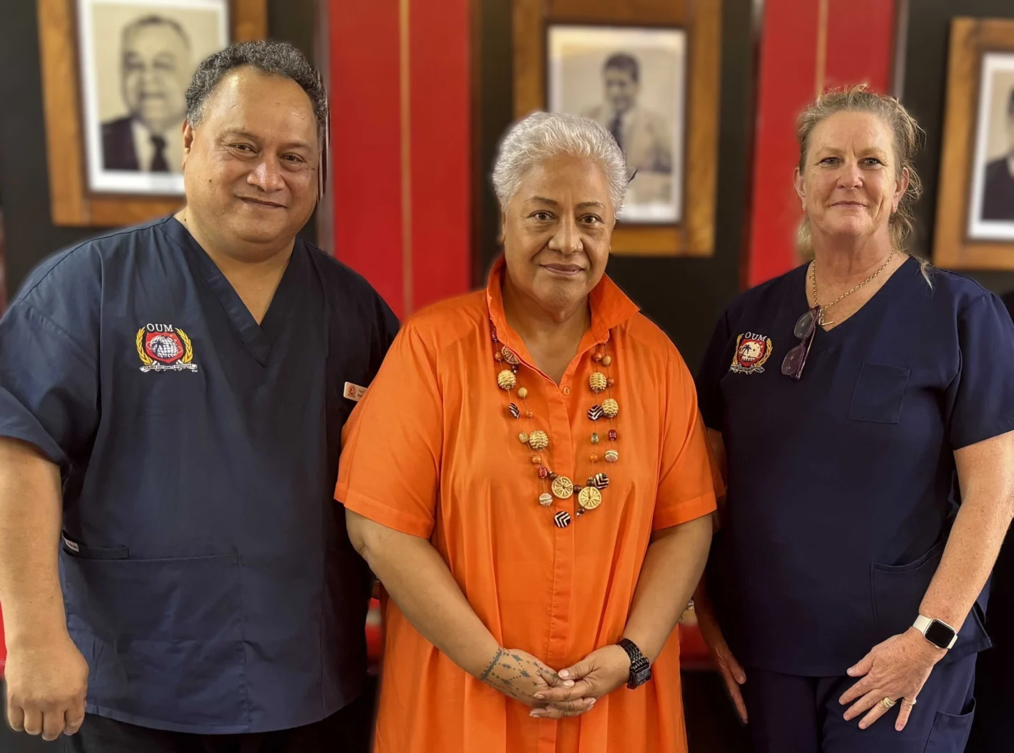 Three smiling people pose for a photo. A man wearing a dark blue scrubs. A woman wearing a vibrant orange dress. To her right is a woman in a dark blue scrubs.