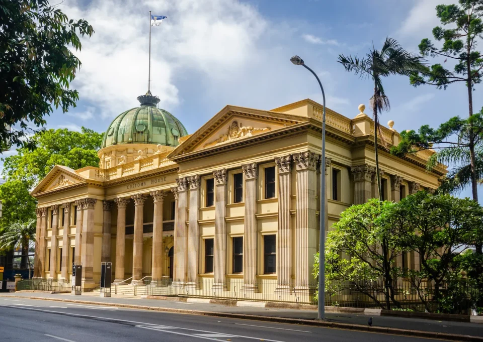 A grand, neoclassical building with a golden dome and colonnades, set on a tree-lined street.