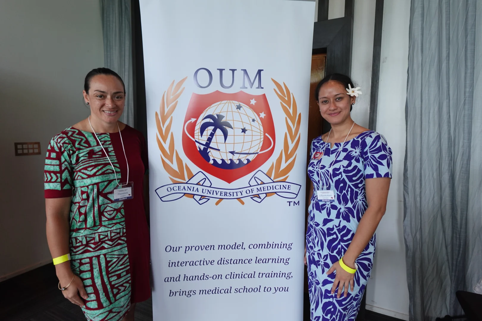 Two smiling women in blue floral dresses stand beside a banner for 'Oceania University of Medicine' and 'OUM'. Banner reads, "Our proven model, combining interactive distance learning and hands-on clinical training, brings medical school to you."