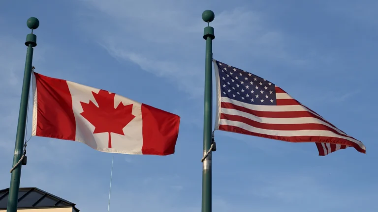 Canadian flag and United States flag side by side on flagpoles against a blue sky