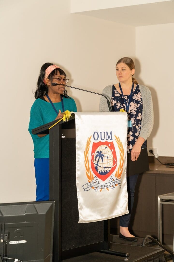 Two women on stage one talking one watching her OUM podium banner