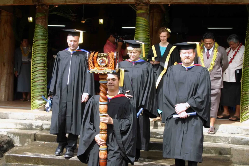 A group of four people outdoors, graduates and faculty, in black academic gowns and mortarboards stand on a stone staircase at a graduation ceremony.