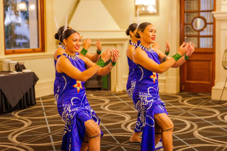 Group of women performing a traditional Samoan dance indoors, wearing blue floral dresses with Polynesian patterns, green wristbands, shell necklaces, and decorative hair ornaments.