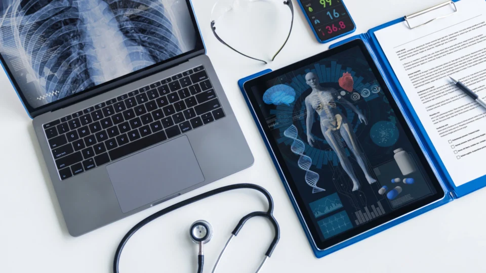 Laptop showing chest X-ray, tablet displaying human anatomy and DNA graphics, stethoscope, medical chart with pen, and digital vitals monitor on a desk, representing evidence-based medical study and practice