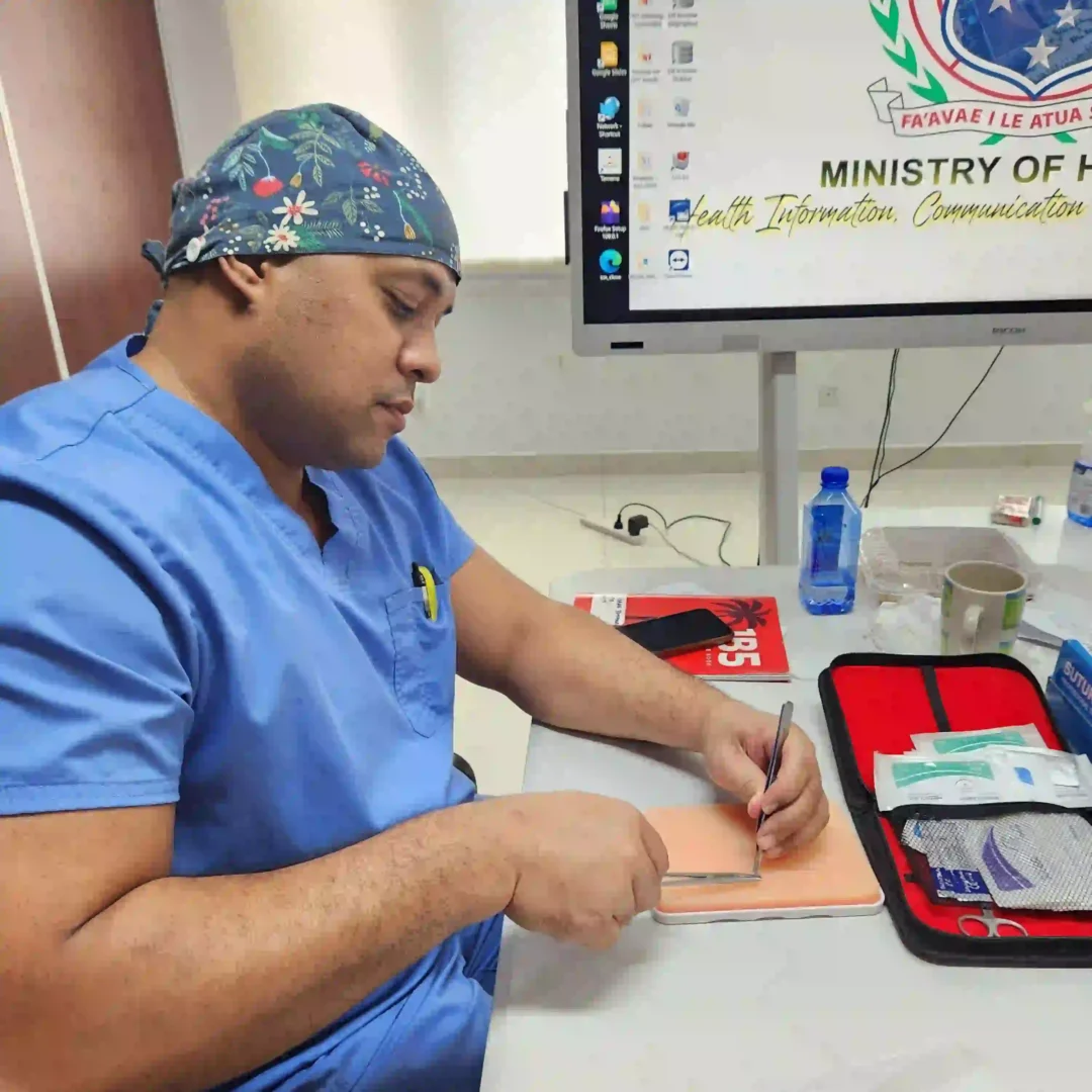 A man wearing a light green surgical hat with a floral design is seated at a desk. He is looking down at an open, red plastic box filled with medical supplies, including syringes and bandages.
