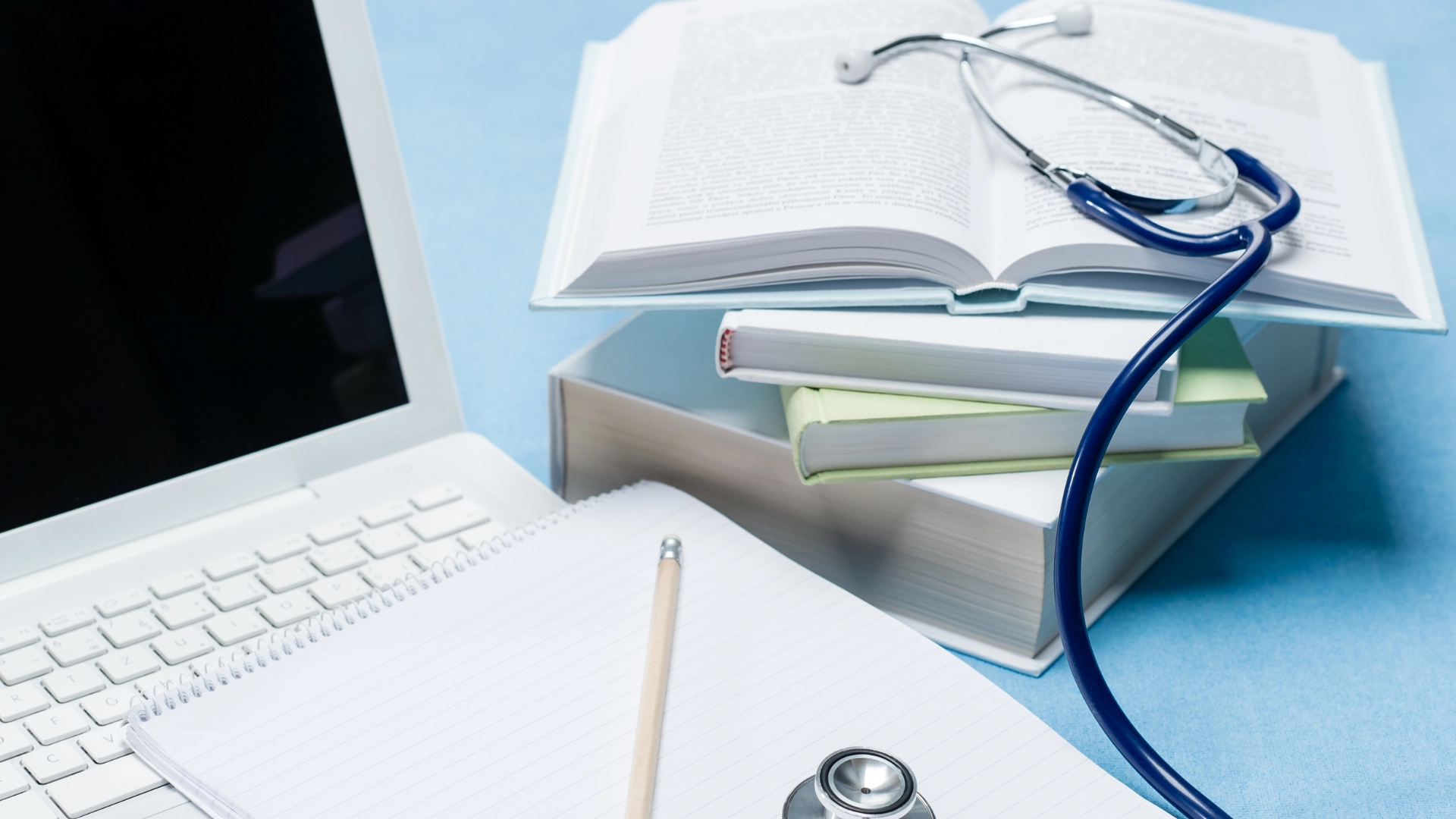 Stack of medical textbooks with a stethoscope on top, next to a laptop and notebook with pencil, representing online medical education and study