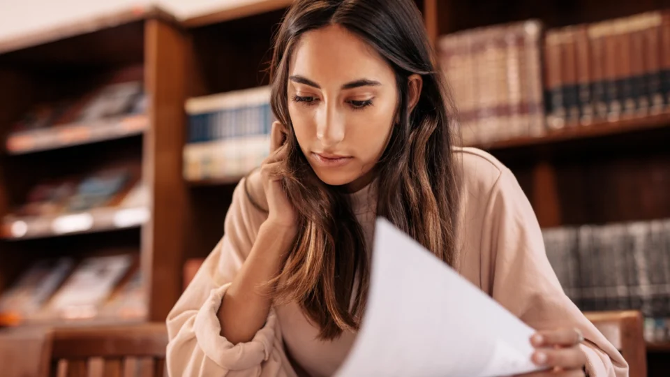 A young woman with long dark hair is sitting in front of bookshelves, intently reading a document.