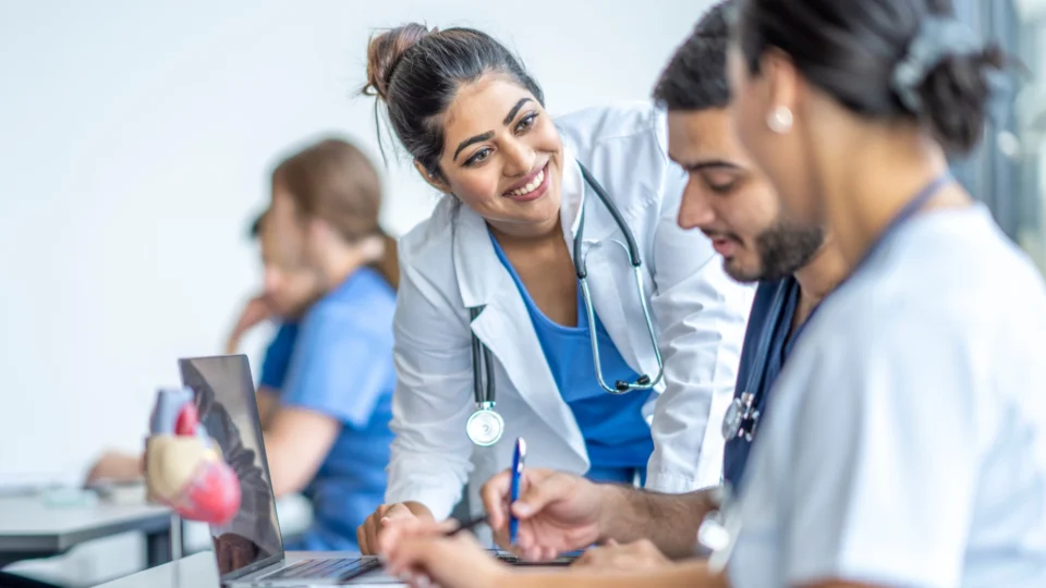 A smiling female doctor in white jacket and blue scrubs is explaining something to a male medical student, who is looking at a laptop.