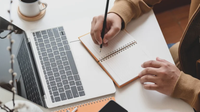 Close-up of a person writing in a spiral notebook while working on a laptop at a desk, with a coffee cup and smartphone nearby