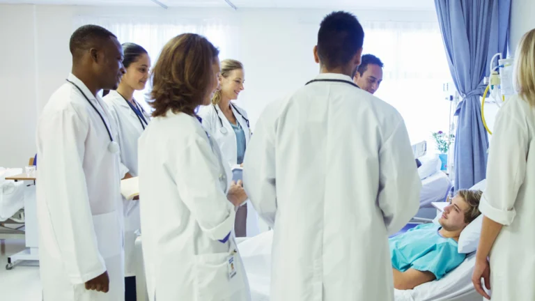 Group of medical students and doctors in white coats conducting bedside teaching rounds with a patient lying in a hospital bed