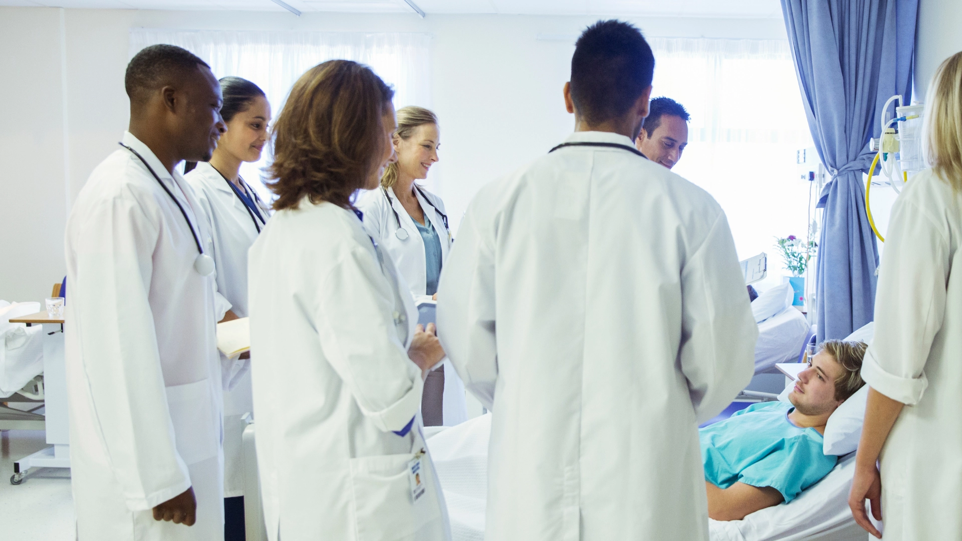 Group of medical students and doctors in white coats conducting bedside teaching rounds with a patient lying in a hospital bed