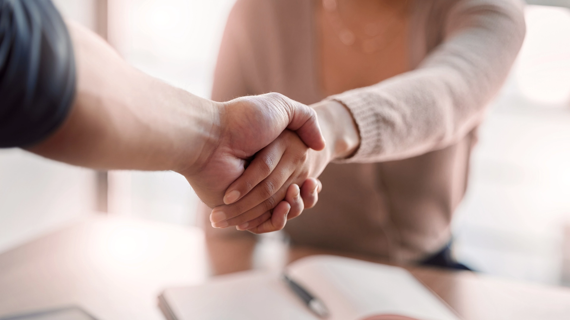 Close-up of two people shaking hands across a desk, symbolizing agreement or partnership, with a pen and notebook visible in the blurred background