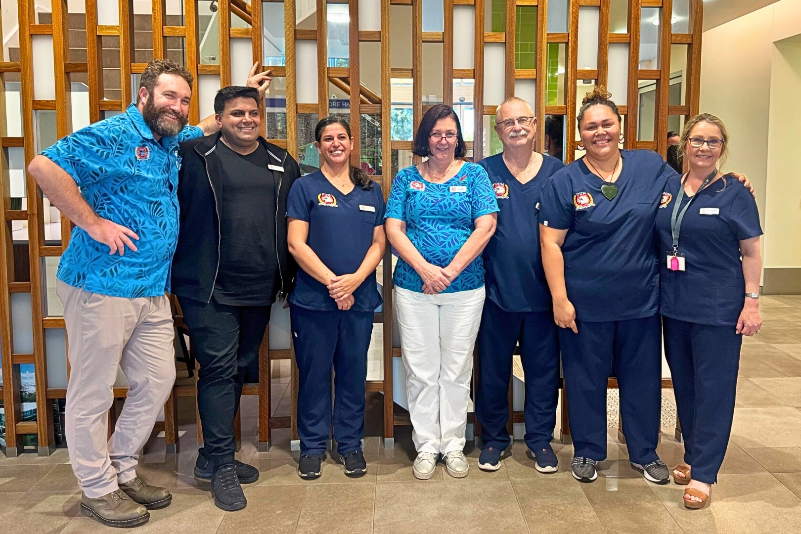 Seven healthcare professionals, men and women of various ethnicities, pose for a photo standing in a hospital hallway. Five of the individuals are wearing dark blue scrubs, while two are wearing different colored shirts.