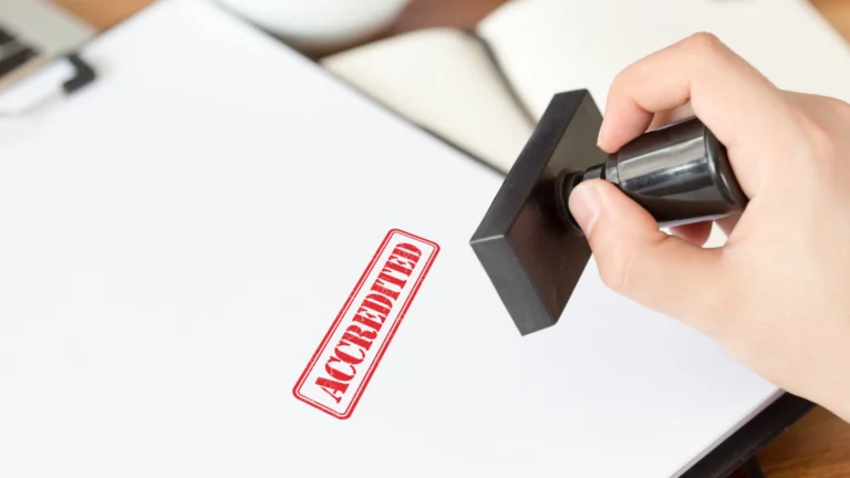 Close-up of a hand pressing a stamp that leaves a red “ACCREDITED” mark on a white sheet of paper