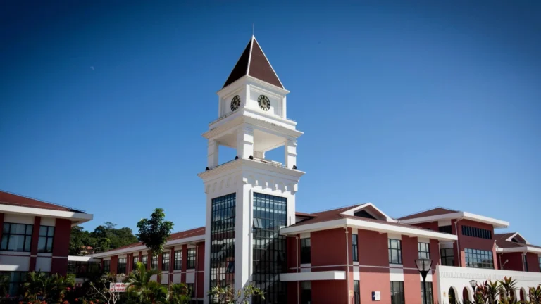 Large red and white building with a tall clock tower and glass windows, set against a clear blue sky in Samoa