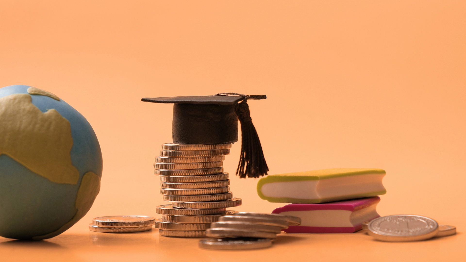 Stack of coins topped with a graduation cap beside miniature books and a globe, symbolizing education costs, scholarships, and global learning opportunities
