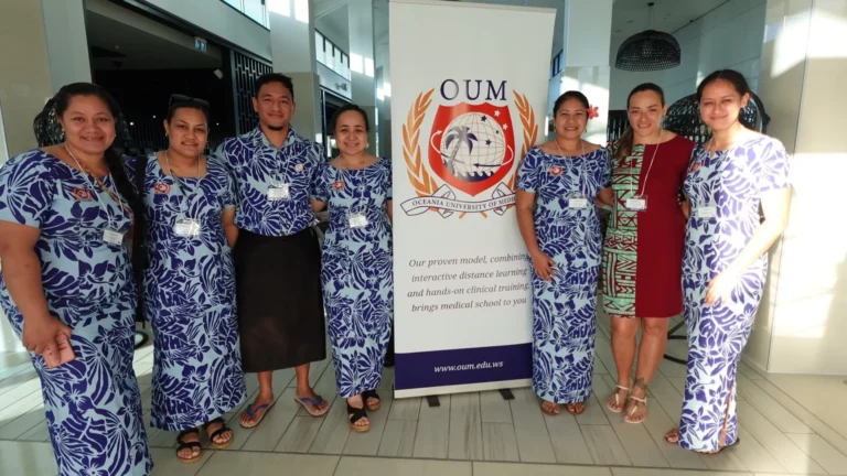 Group of men and women in traditional Samoan attire standing beside an Oceania University of Medicine banner at a medical conference
