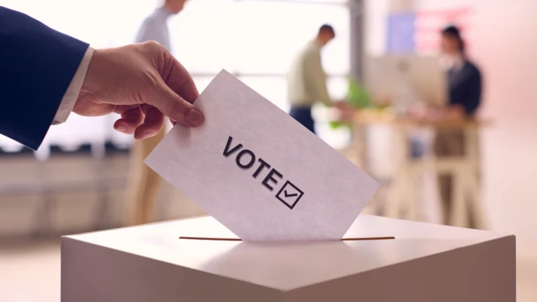 Close-up of a person placing a ballot labeled “VOTE” with a checkmark into a ballot box, with blurred figures in the background