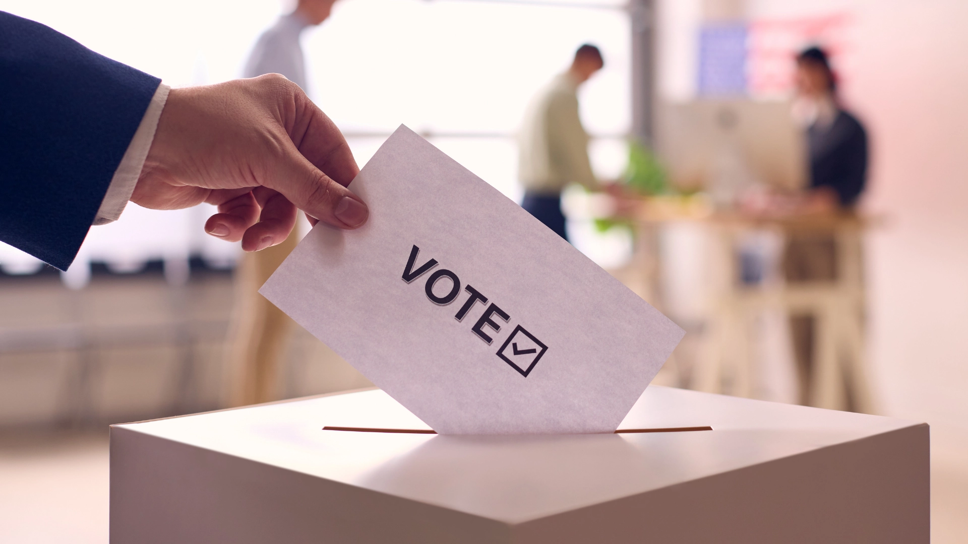 Close-up of a person placing a ballot labeled “VOTE” with a checkmark into a ballot box, with blurred figures in the background