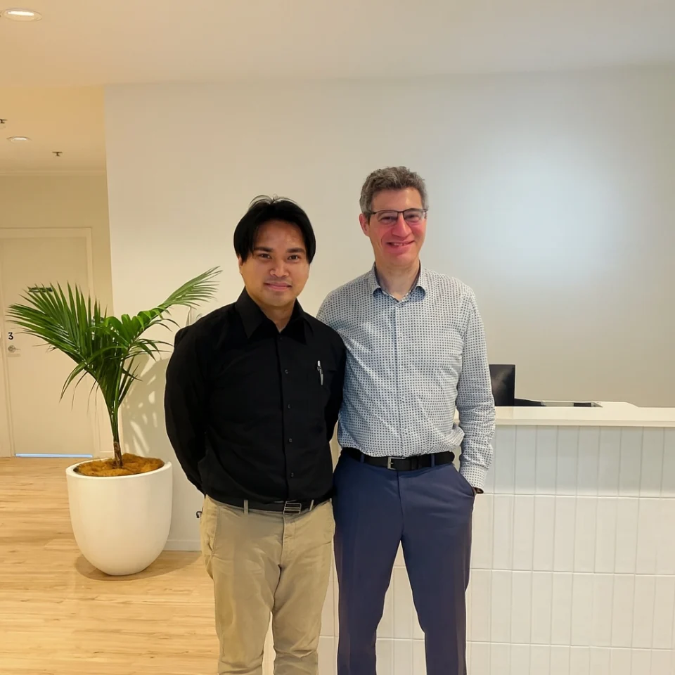 Two men posing for a photo, one with dark hair and the other with graying hair, standing in what appears to be an office lobby.