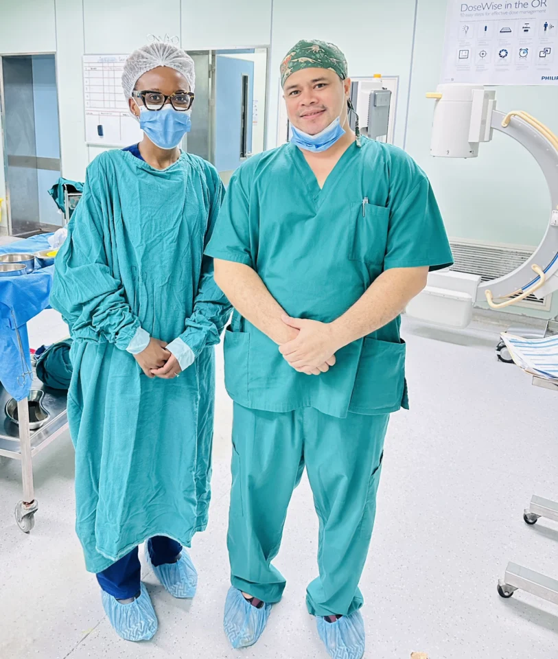 Three medical professionals in teal scrubs and blue shoe covers pose for a photo in an operating room setting.