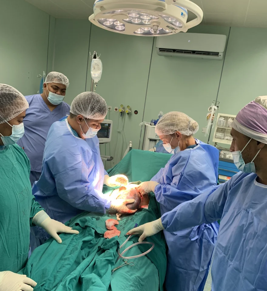 A surgical team is operating on a patient in an operating room. Several masked and gowned medical professionals are gathered around the operating table.
