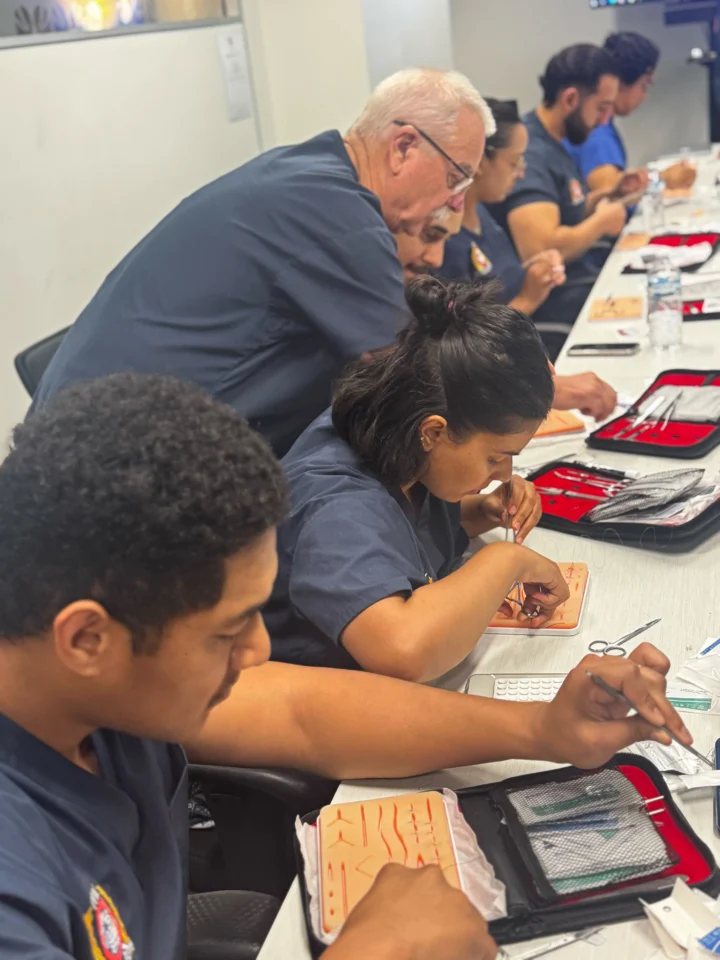 A group of medical students wearing dark blue scrubs working at desks in a classroom, with an instructor leaning over to assist them with their work.