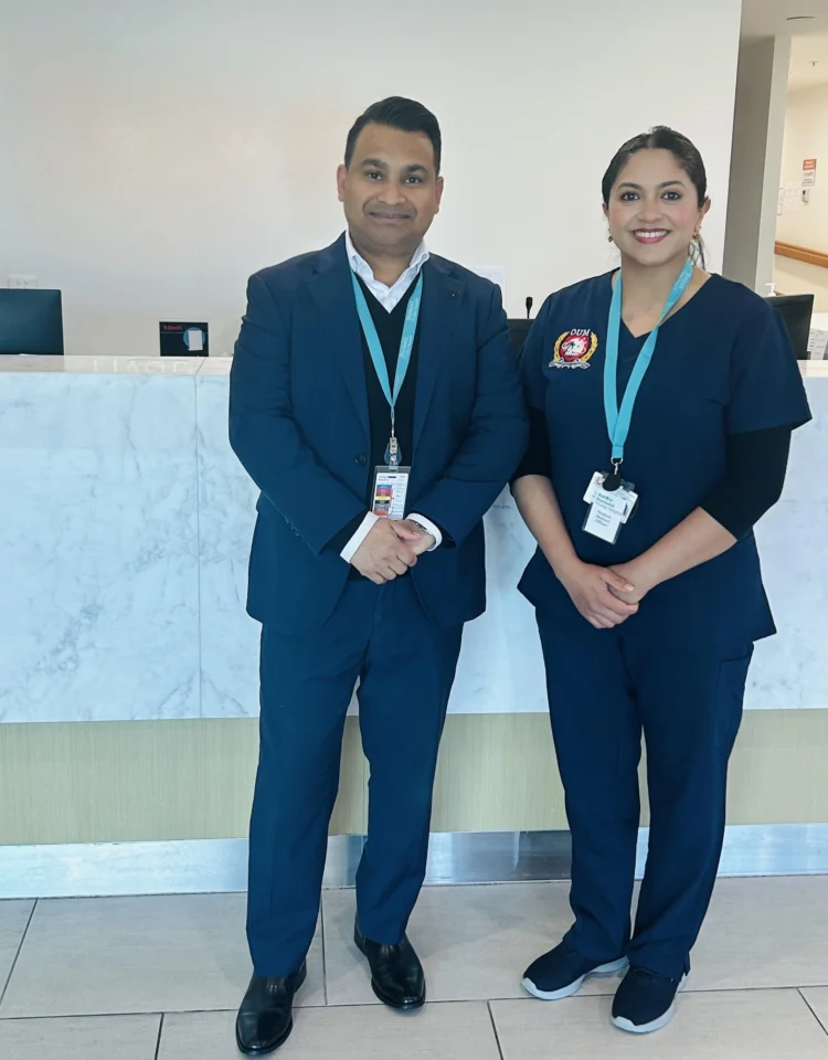 Two healthcare workers, a man and a woman, stand side-by-side in a hospital setting. They are both wearing blue scrub and name badges
