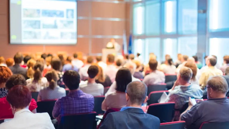 Audience seated in a conference room listening to a speaker at a podium with a presentation projected on a large screen