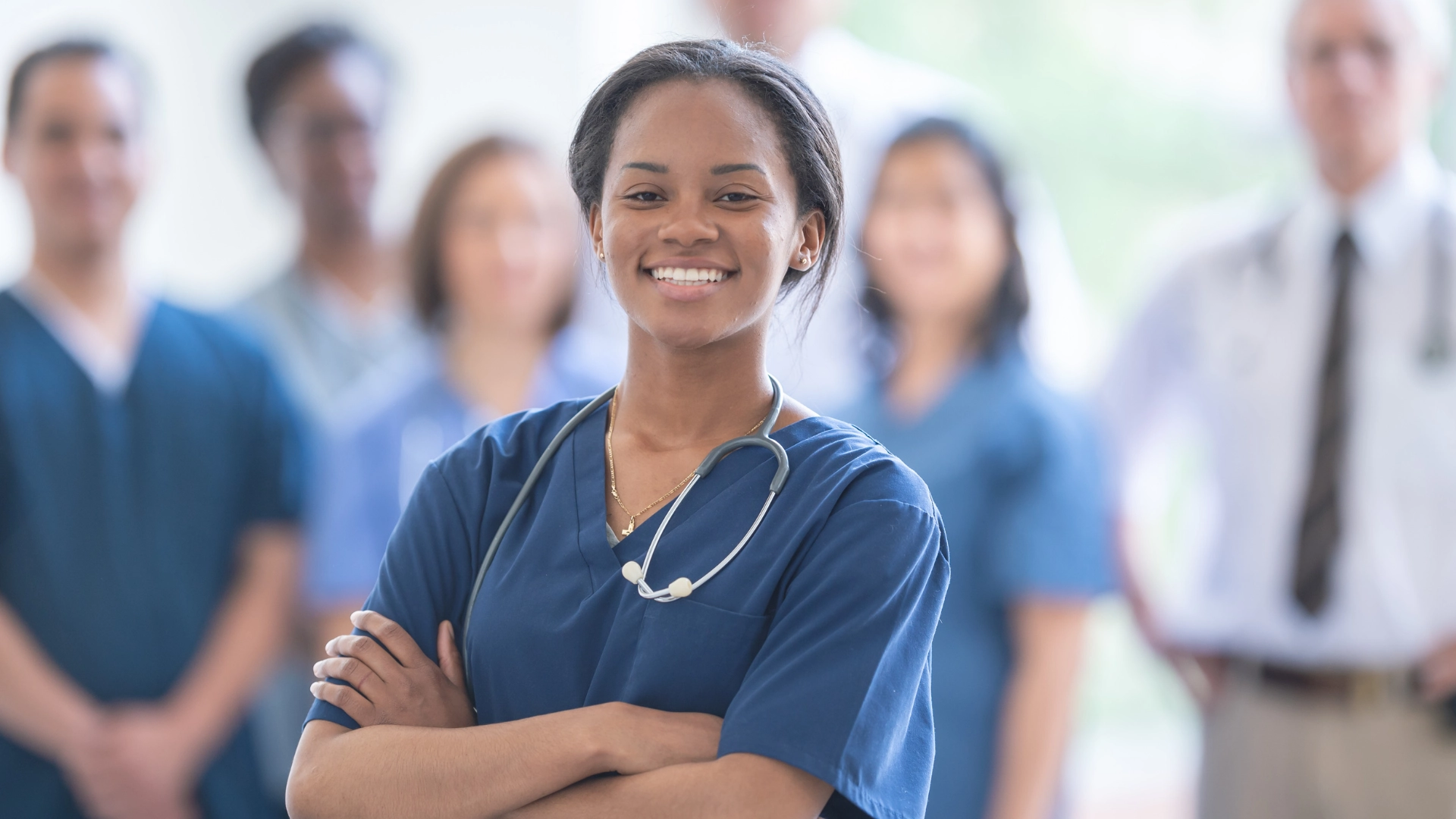 Smiling medical professional in blue scrubs with a stethoscope around her neck standing confidently with arms crossed, with a blurred group of healthcare workers in the background