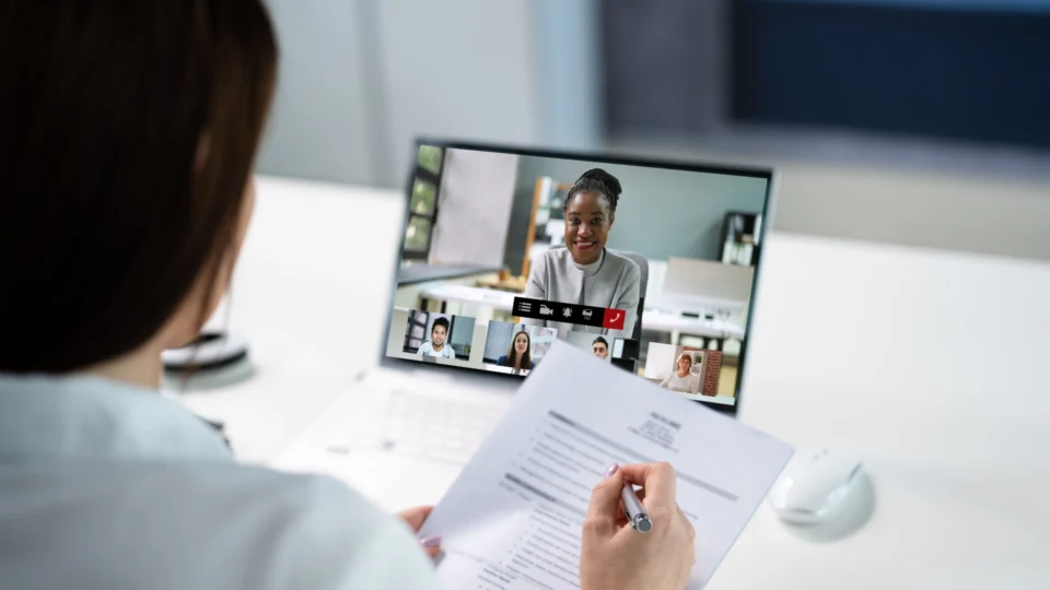 Person holding interview notes while participating in a virtual video interview with multiple participants displayed on a laptop screen