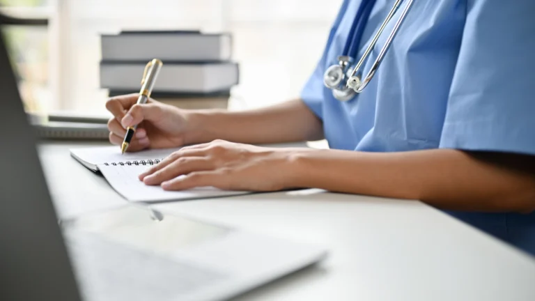 Medical student in blue scrubs with a stethoscope around their neck writing notes in a notebook at a desk with a laptop and stack of books